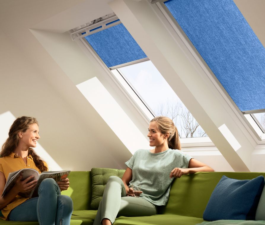 Zwei Frauen auf einer Couch unterhalten sich, mit zwei VELUX Dachfenstern mit zwei blauen VELUX Hitzeschutz-Markisen im Hintergrund.