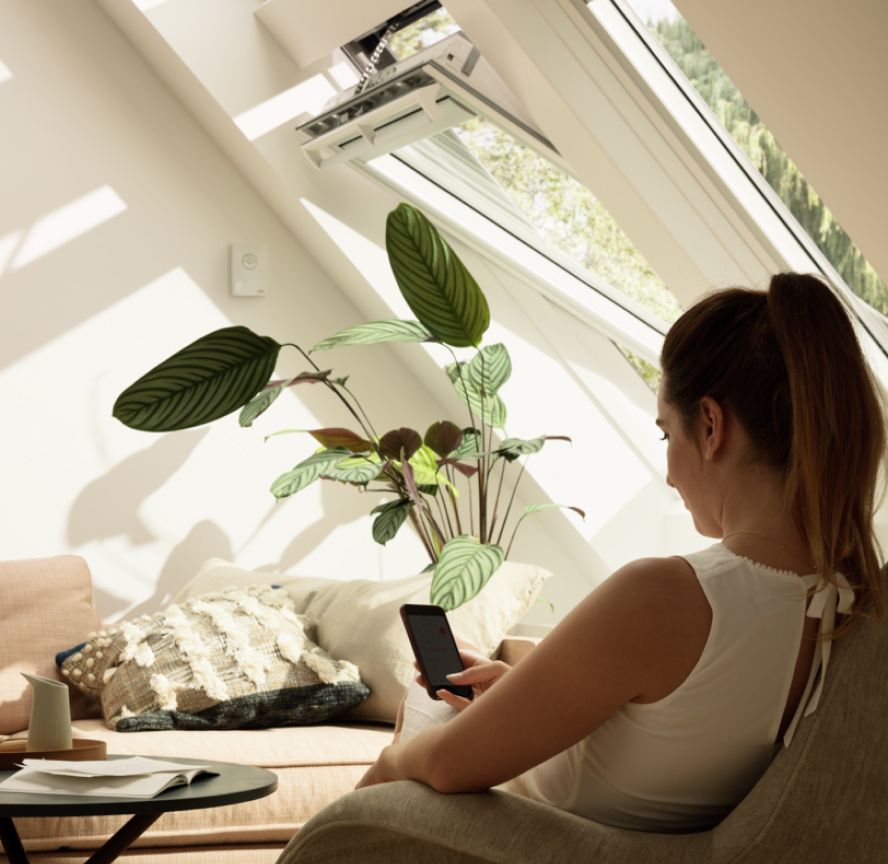 Donna che controlla le sue finestre per tetti VELUX nel soggiorno con l'app VELUX ACTIVE sul suo smartphone.