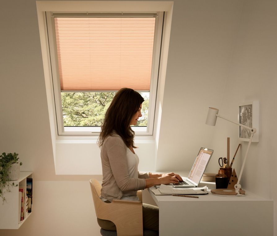 Femme assise dans son espace de télétravail moderne écrivant un e-mail sur son ordinateur portable avec une fenêtre de toit VELUX derrière elle, munie d'une marquisette beige.