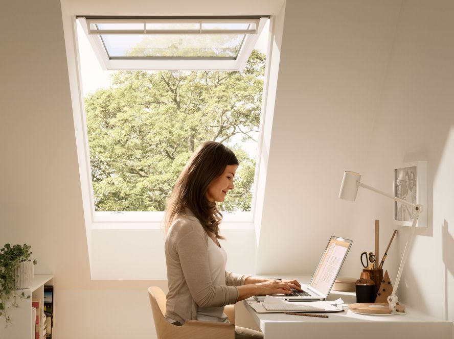 Frau in ihrem modernen Homeoffice, beim E-Mail-Schreiben auf ihrem Laptop mit einem VELUX Dachfenster im Hintergrund, das zwecks Frischluftzufuhr geöffnet ist.