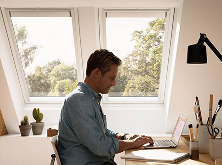 Ein Mann, an seinem Schreibtisch arbeitend, natürliches Licht strömt durch zwei gestapelte VELUX Dachfenster. 
