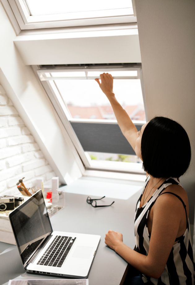 Une femme est assise à un bureau avec son ordinateur et tend la main pour ouvrir sa fenêtre de toit VELUX.