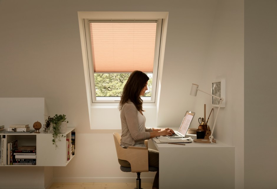 Une femme travaille à son bureau; une fenêtre de toit VELUX est derrière elle et elle a un store coloré abaissé.
